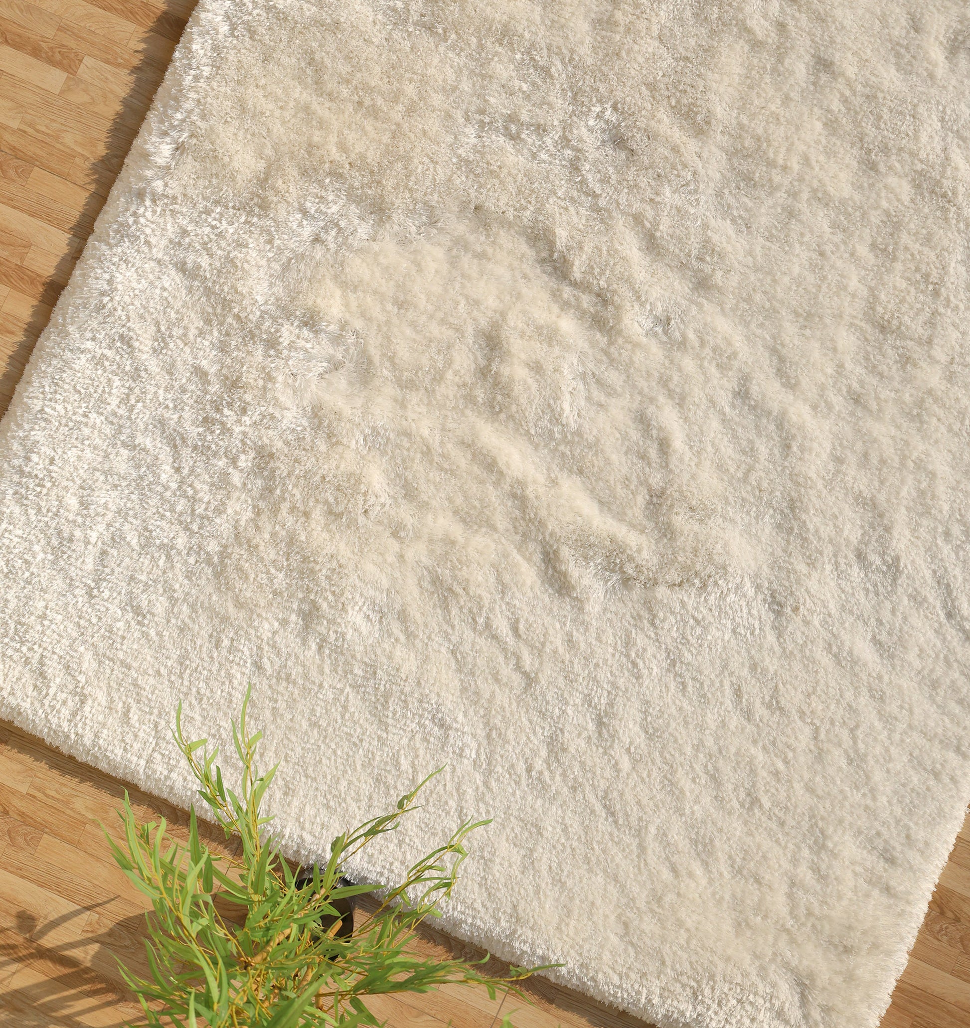 The RAMSHA HOME White Color Plush Shaggy Area Rug - SG 040 features plush fibers that add cozy warmth to a light wooden floor, with a green potted plant partially visible in the bottom left corner.