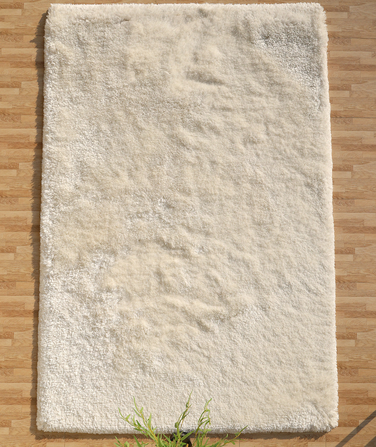 The RAMSHA HOME White Color Plush Shaggy Area Rug - SG 040, featuring dense off-white fibers, rests on a light wooden floor with its edge bordered by a green leafy plant for a cozy touch.