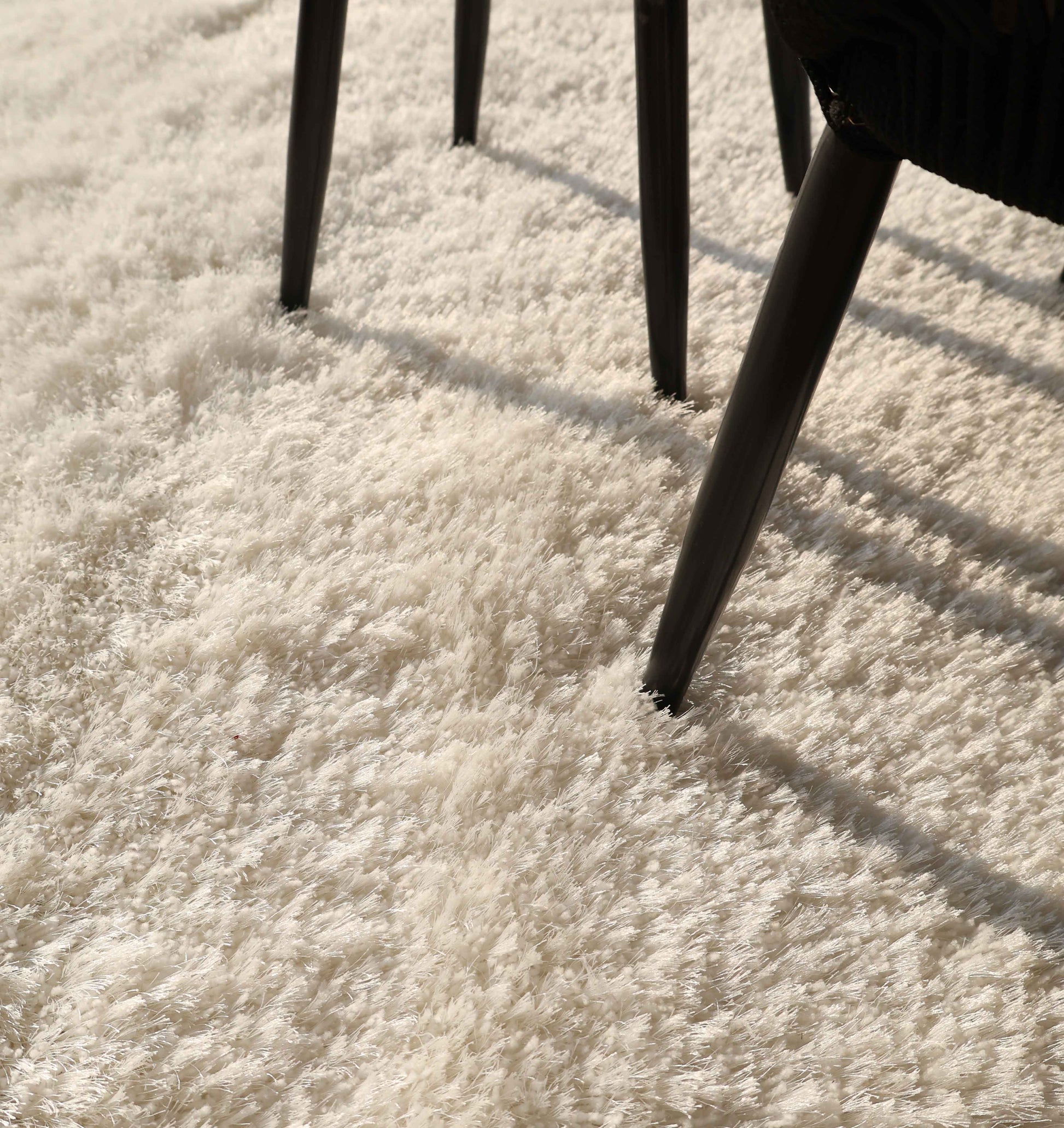 Close-up of a chair's legs casting shadows on the RAMSHA HOME White Color Plush Shaggy Area Rug - SG 040, highlighting its thick plush fibers and creating a cozy, sunlit atmosphere.
