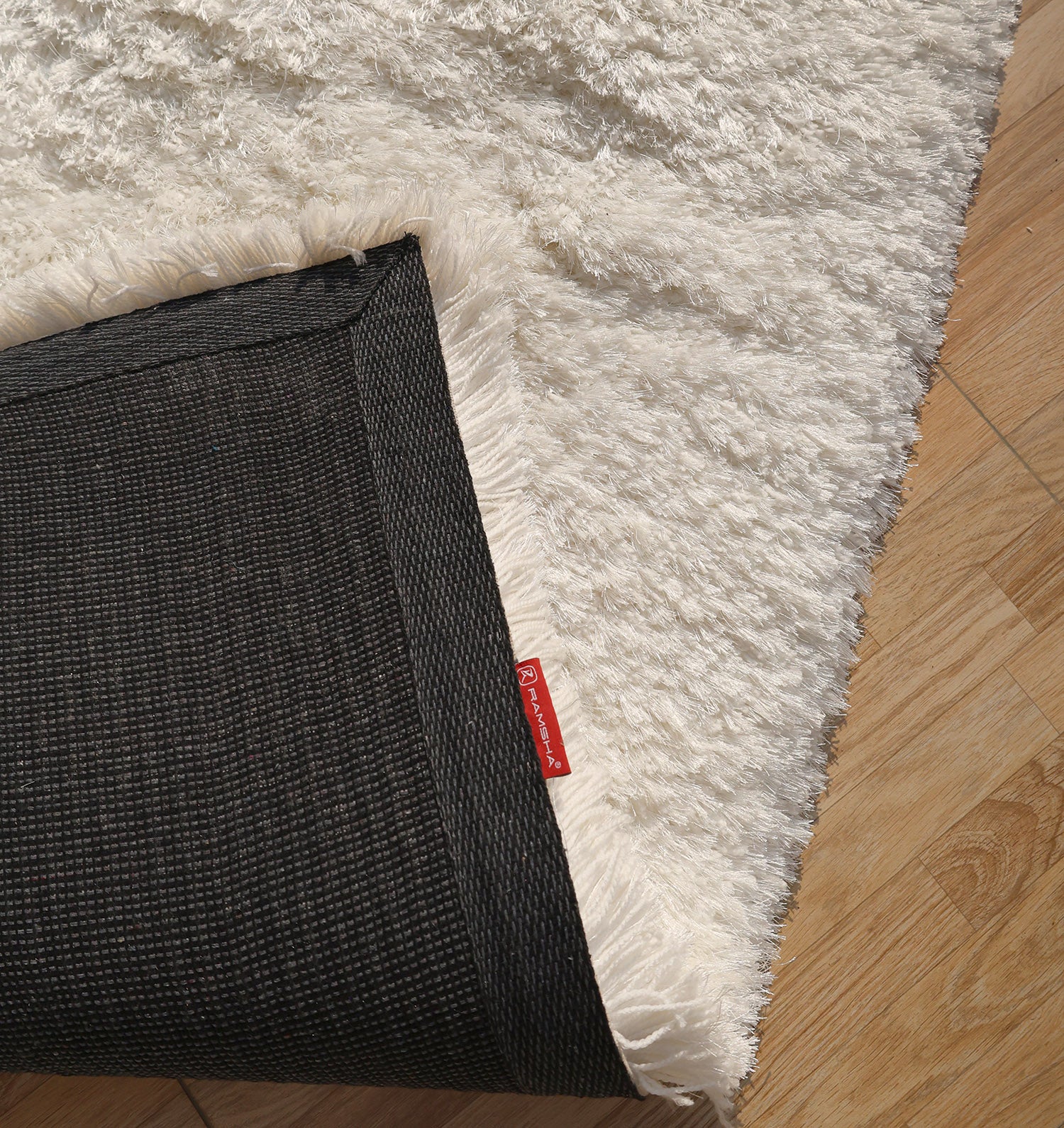 The RAMSHA HOME White Color Plush Shaggy Area Rug - SG 040 lies on a wooden floor with a flipped corner revealing its plush fibers, black non-slip backing, and red tag—perfect for adding cozy style to any room.