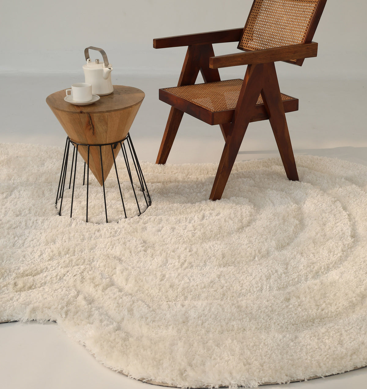A wooden chair with a woven seat and a small table holding a teapot and cups are arranged atop the RAMSHA HOME White Irregular Shaped Home Decor Shaggy Rug (SG 040 3D), adding texture and warmth to the minimalist setting.