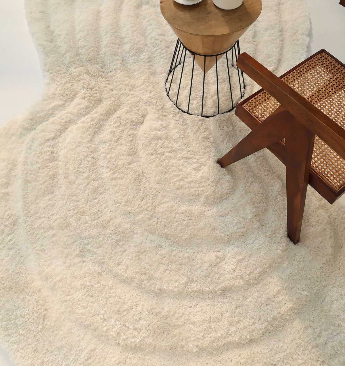 The RAMSHA HOME White Irregular Shaped Home Decor Shaggy Rug - SG 040 3D is featured in a minimal and cozy scene alongside a wooden chair with a woven cane seat and a small round side table displaying two white vases.
