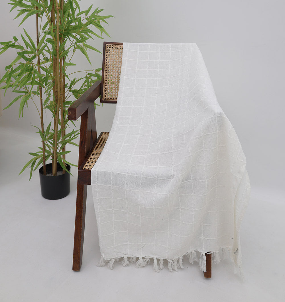 The White Decor Throw TAL 051 (130x160cm) by Ramsha Home, with a subtle grid pattern and white fringe, is draped over a wooden chair with a woven seat next to a potted bamboo plant on a light background.