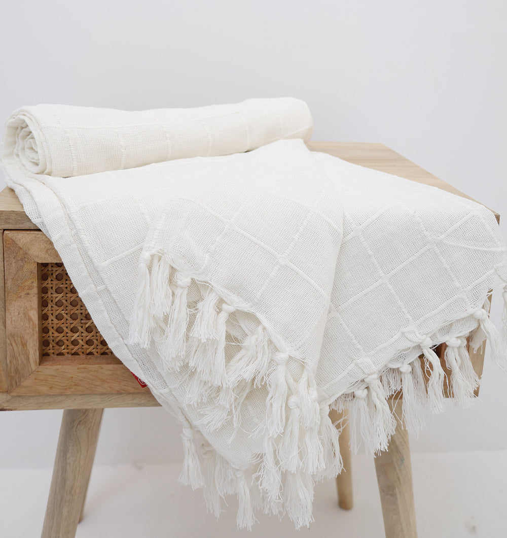 The Ramsha Home White Decor Throw TAL 051 (130x160cm) features a subtle grid pattern and fringe edges, draped over a wooden stool with a woven panel, adding modern comfort to any space against a plain white background.