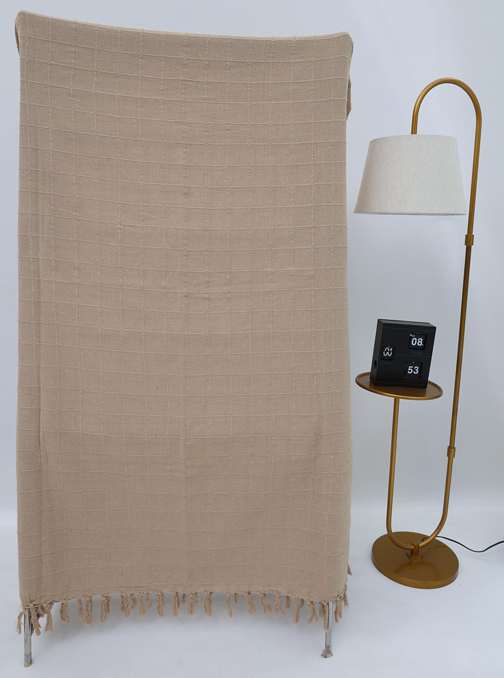 The Ramsha Home Living Room Throw Beige Color TAL 052 (130x160cm) with a grid pattern and fringe hangs over a stand near a gold floor lamp with a round shelf, where a black flip clock shows 08:53 against the white background.