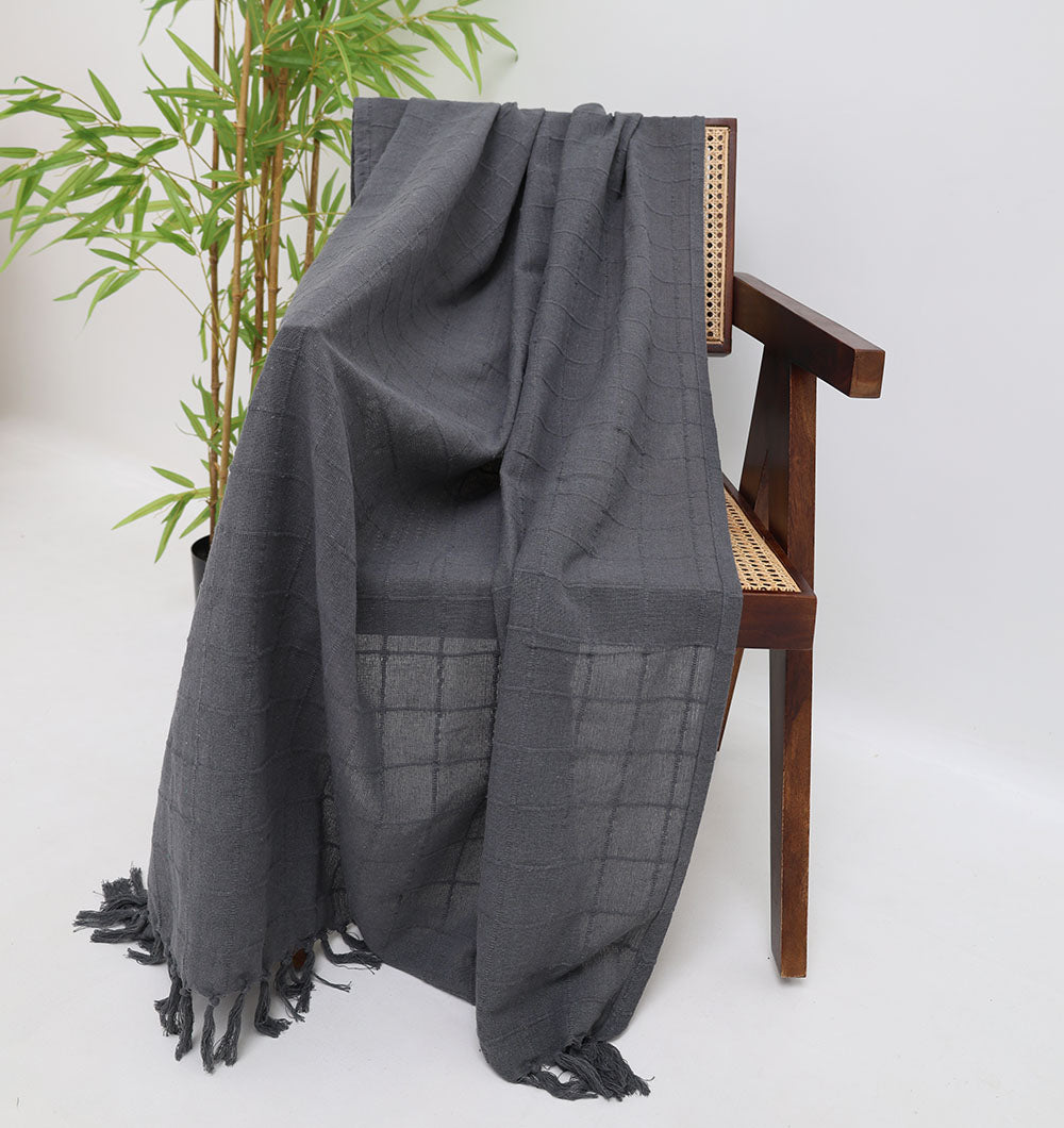 The Ramsha Home Living Room Throw in Dark Grey (TAL 053, 130x160cm) is draped over a wooden chair with a cane seat beside a green potted plant, all set against a plain white background.