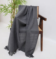 The Ramsha Home Living Room Throw in Dark Grey (TAL 053, 130x160cm) is draped over a wooden chair with a cane seat beside a green potted plant, all set against a plain white background.