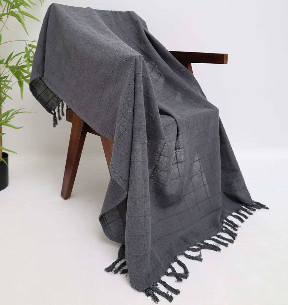The Ramsha Home Living Room ThrowDark Grey Color TAL 053 (130x160cm) is draped over a wooden chair, showcasing its subtle checkered pattern and fringe edges. A leafy plant appears to the left, with a plain white background behind.