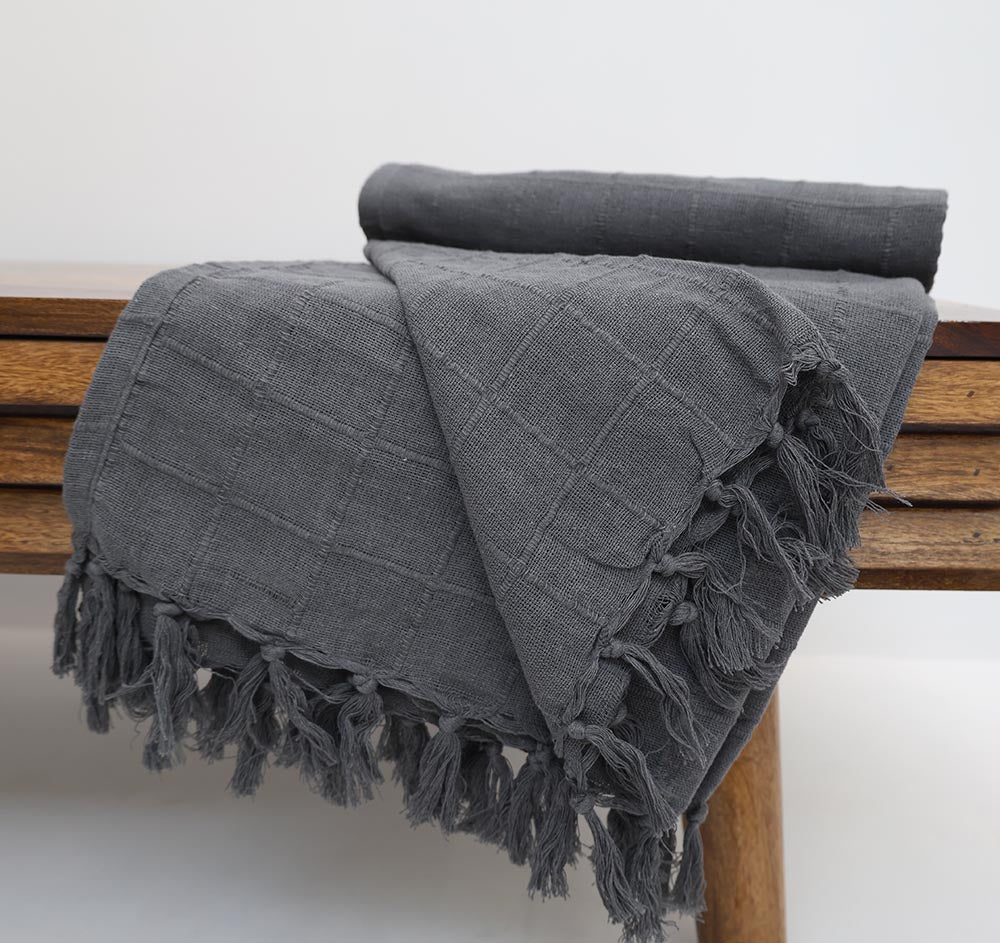 The Ramsha Home Living Room ThrowDark Grey Color TAL 053 (130x160cm) is draped over a wooden bench, showing off its fringe edges against a plain white background.