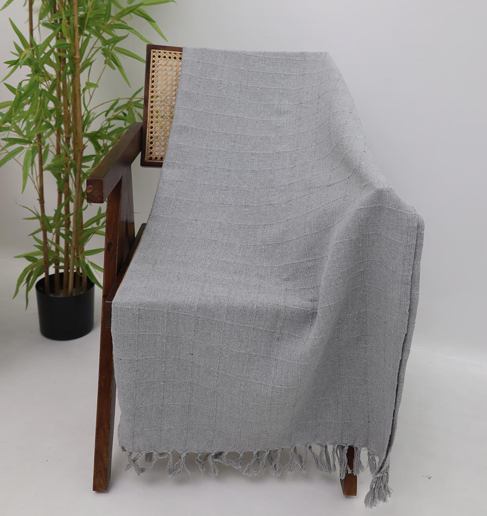 The Living Room Throw Light Grey Color TAL 054 (130x160cm) by Ramsha Home is draped over a wooden chair with a woven backrest, accenting a cozy living room with its textured cotton and fringed edges. A potted plant sits nearby on the white floor.