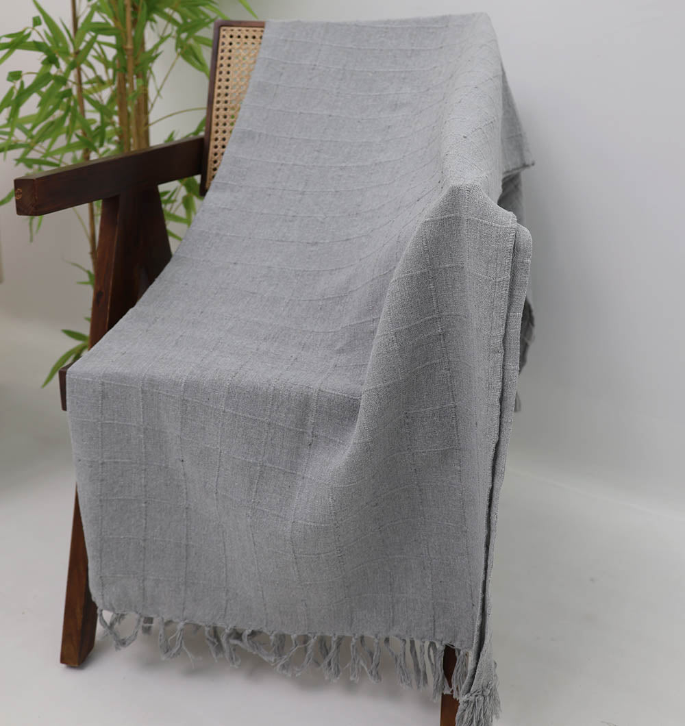 The Ramsha Home Living Room Throw Light Grey Color TAL 054 (130x160cm) adds style draped on a wooden chair with woven cane backrest, set against a white wall and green plant. Perfect for elevating your living room décor.