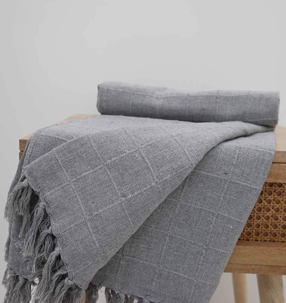 The Living Room Throw Light Grey Color TAL 054 (130x160cm) by Ramsha Home is draped over a wooden stool with a cane panel, adding cozy luxury to any living room decor against a plain white background.