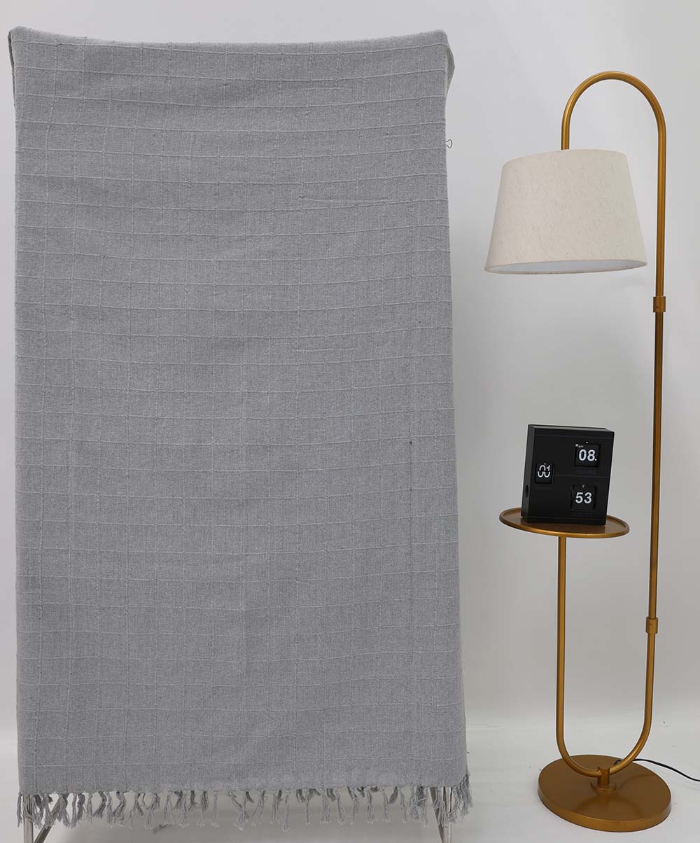 The Living Room Throw Light Grey Color TAL 054 (130x160cm) by Ramsha Home: Luxury Rugs & Home Furnishings Brand In The UAE hangs near a gold floor lamp with a round shelf and black digital clock, creating stylish modern decor against a white backdrop.