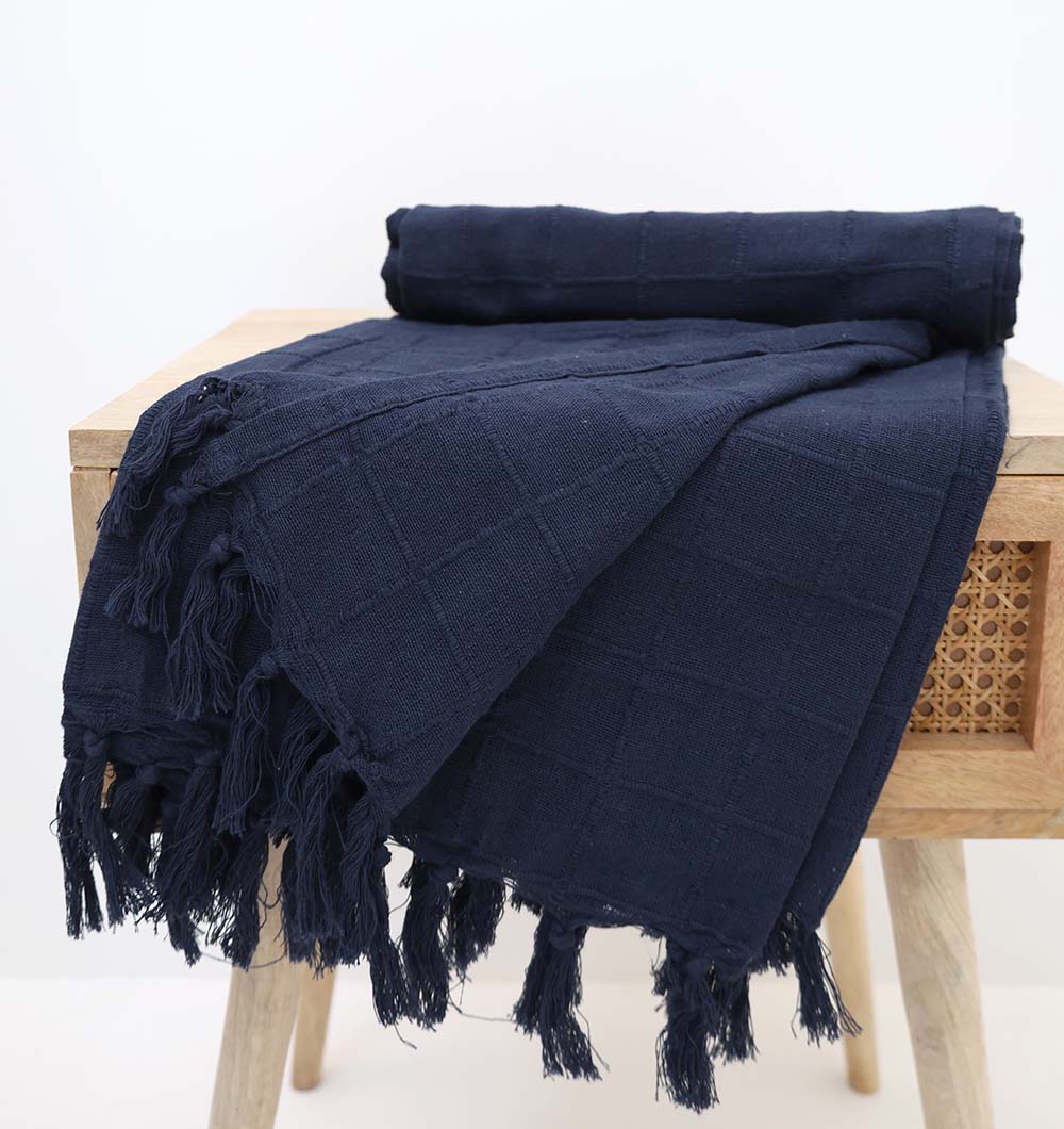 The Ramsha Home Living Room Throw Navy Blue Color TAL 055 (130x160cm), draped over a light wooden stool with fringed edges and a rolled section, adds an elegant, cozy touch to your living room decor.