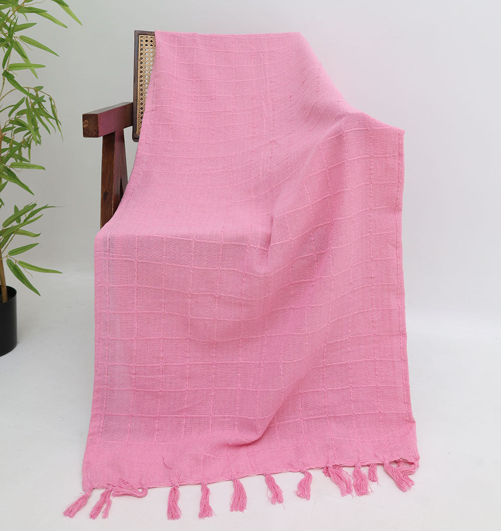 A wooden chair displays the Living Room Throw Pink Color TAL 056 (130x160cm) by Ramsha Home, featuring a subtle grid pattern and tasseled edges. A green potted plant on the left brings freshness to your living room decor.
