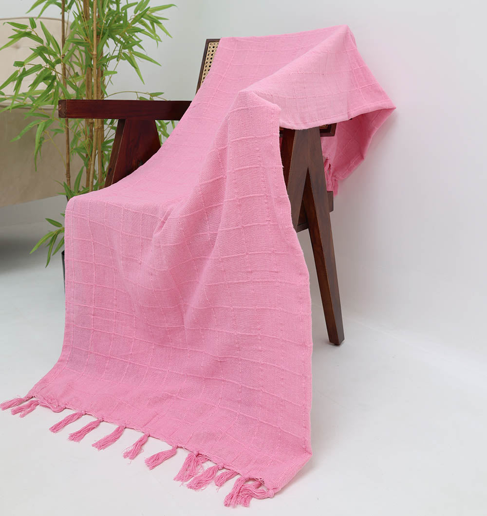 The Ramsha Home Living Room Throw Pink Color TAL 056 (130x160cm), featuring a textured design and tasseled edges, is draped over a dark wooden chair in a bright room with a green potted plant in the background.