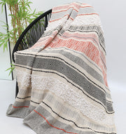 The Living Room Throw Multi Color TAL 058 (130x160 cm) by Ramsha Home features black, white, grey, and orange geometric and striped patterns, draped over a black chair beside a green plant against a plain white background.