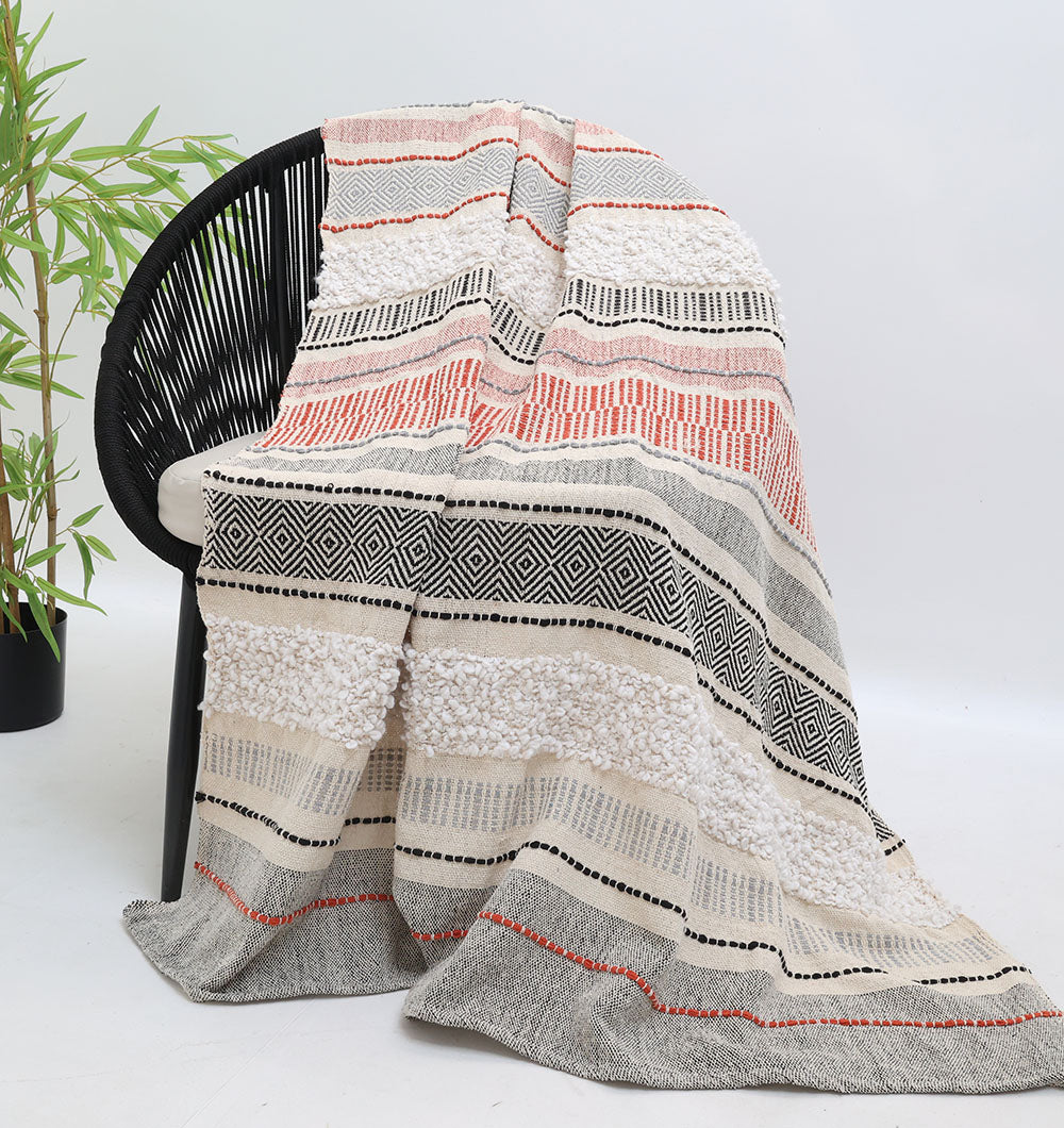 A black chair is adorned with the Living Room Throw Multi Color TAL 058 (130x160 cm) from Ramsha Home: Luxury Rugs & Home Furnishings Brand In The UAE, featuring white, grey, black, and rust stripes beside a leafy plant on a plain white background.