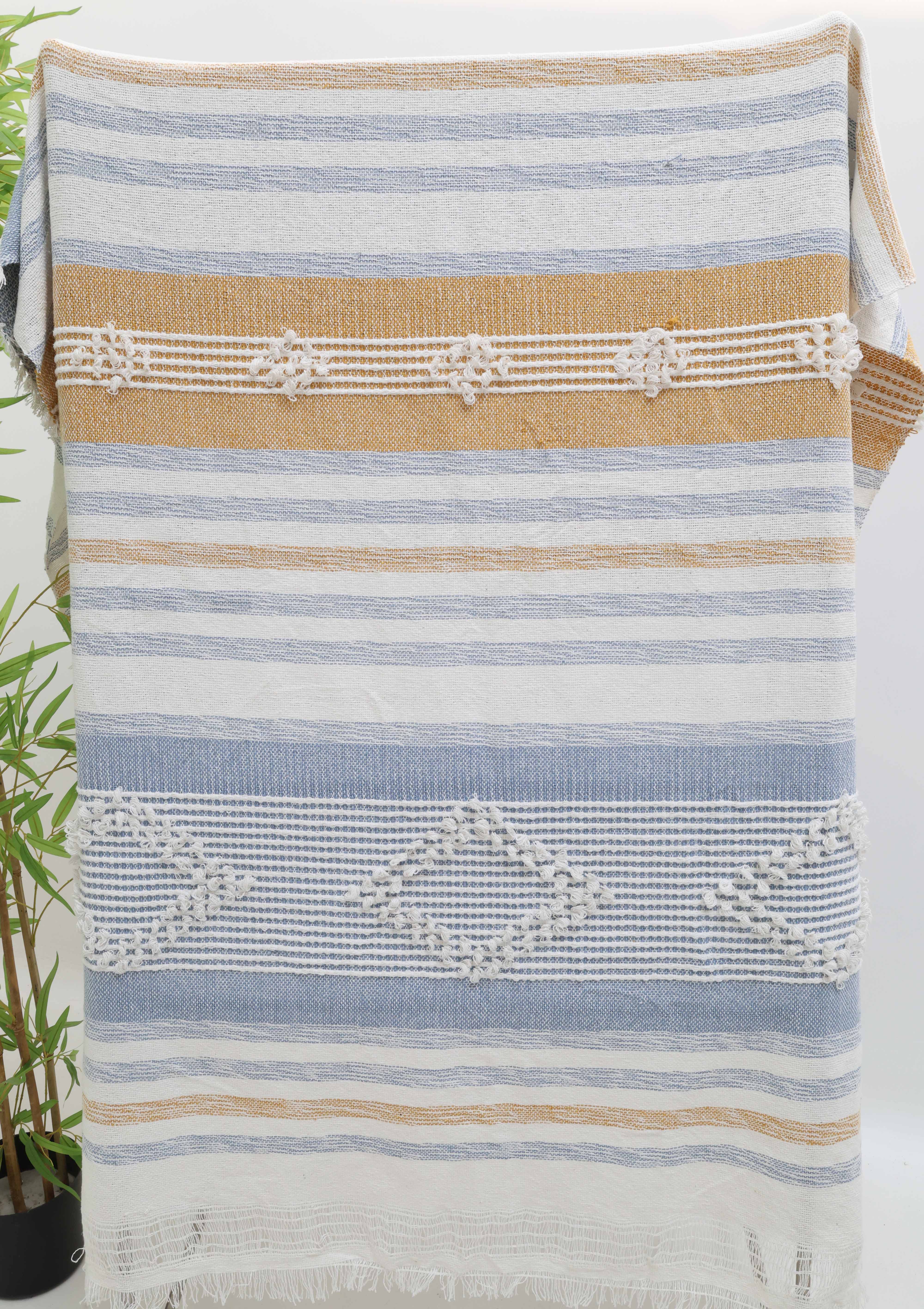 The Living Room Throw Multi Color Cotton Throws TAL 059 (130x150 cm) by Ramsha Home features blue, white, and tan stripes with textured geometric patterns in a soft cotton-acrylic blend, stylishly draped with a leafy green plant on the left.