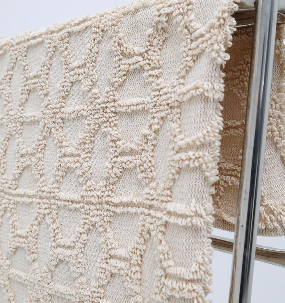 The Living Room Throw Natural Color Cotton Throws TAL 063 (130x175 cm) by Ramsha Home features a textured geometric diamond pattern, adding subtle style to your decor when draped over a rack against a plain background.