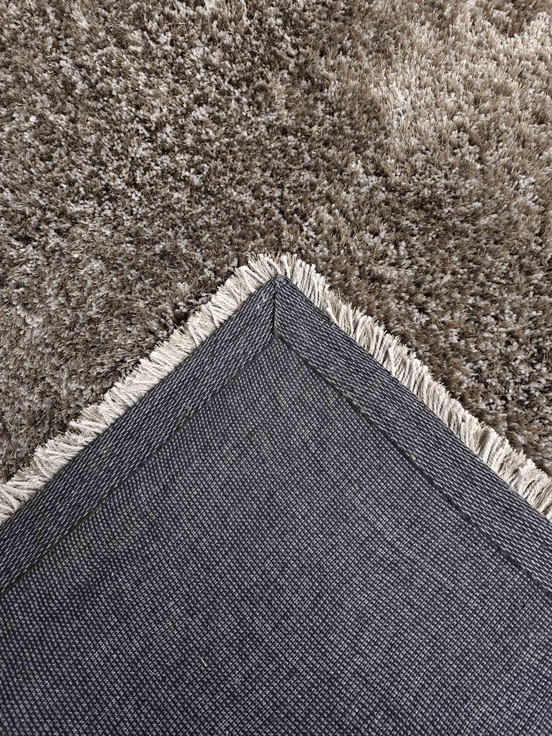A close-up of the RAMSHA HOME Grey Color Rectangle Shaped Shaggy Rug - SG 05, featuring white fringe and a textured backside, displayed atop a soft brown shaggy rug.