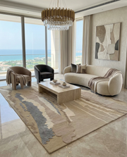 A modern living room with large windows and an ocean view, featuring a beige curved sofa, two armchairs, a rectangular coffee table, gold chandelier, abstract wall art, and the RAMSHA HOME Classic Rectangle Handmade Area Rug-HTWV 177 (300x400 cm) for added elegance.