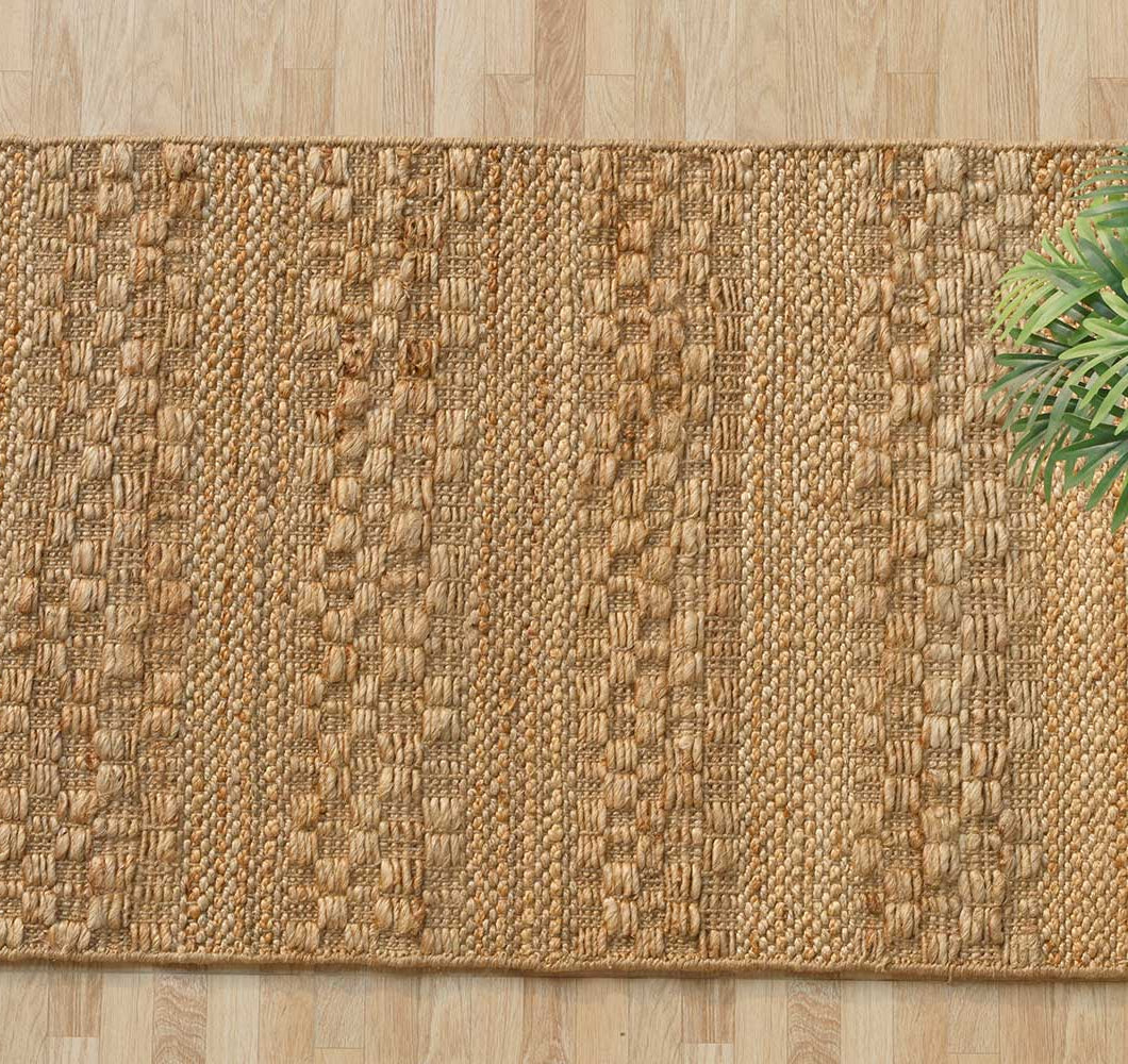 The RAMSHA HOME Natural Color Hand Woven Area Rug WR-034, featuring tan jute and textured stripes, is spread on a light wood floor. A green leafy plant appears in the upper right corner of the image.