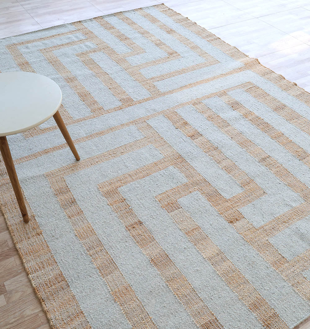 The RAMSHA HOME Jute & Wool Natural Color Hand Woven Rug WR-036 with a beige geometric pattern is displayed on a light wood floor, accompanied by a small round white table with wooden legs in the upper left corner.