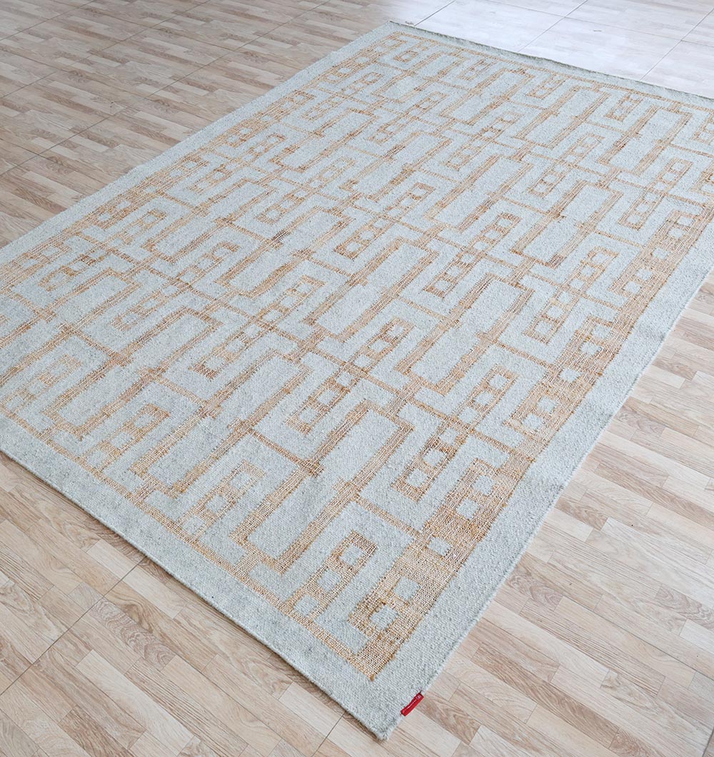The RAMSHA HOME Hand Woven Decor Area Rug WR-037, in light beige and tan with interlocking rectangles and a subtle border, lies on a wood-patterned floor. A small red tag adds detail to this jute wool rug.