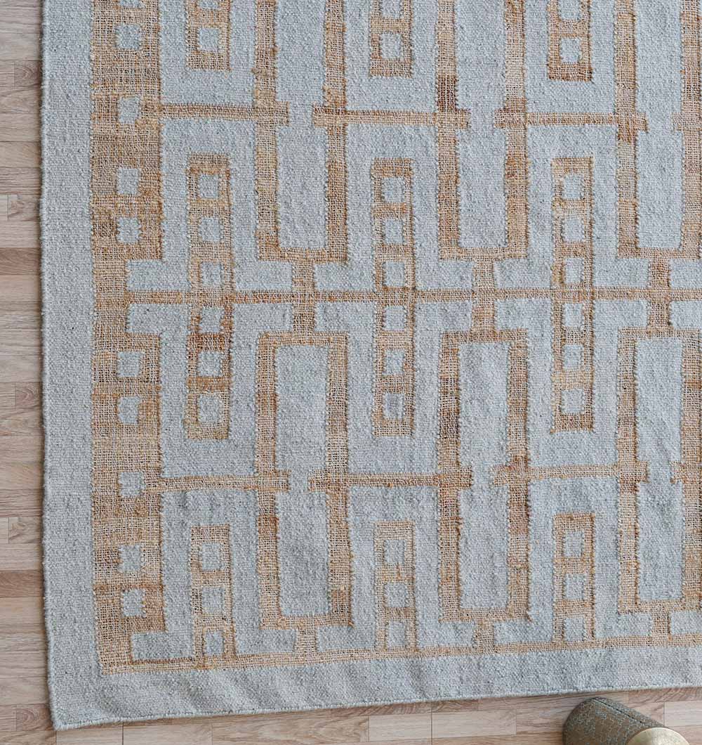 The RAMSHA HOME Hand Woven Decor Area Rug WR-037, featuring a light beige and tan pattern of interlocking lines and rectangles, is laid on a light wood floor with one corner slightly rolled.