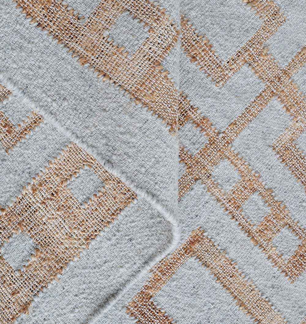 A close-up of the RAMSHA HOME Hand Woven Decor Area Rug WR-037 highlights beige and tan geometric shapes on a light gray base, creating a layered look. The slightly folded fabric emulates the texture of a jute wool rug.
