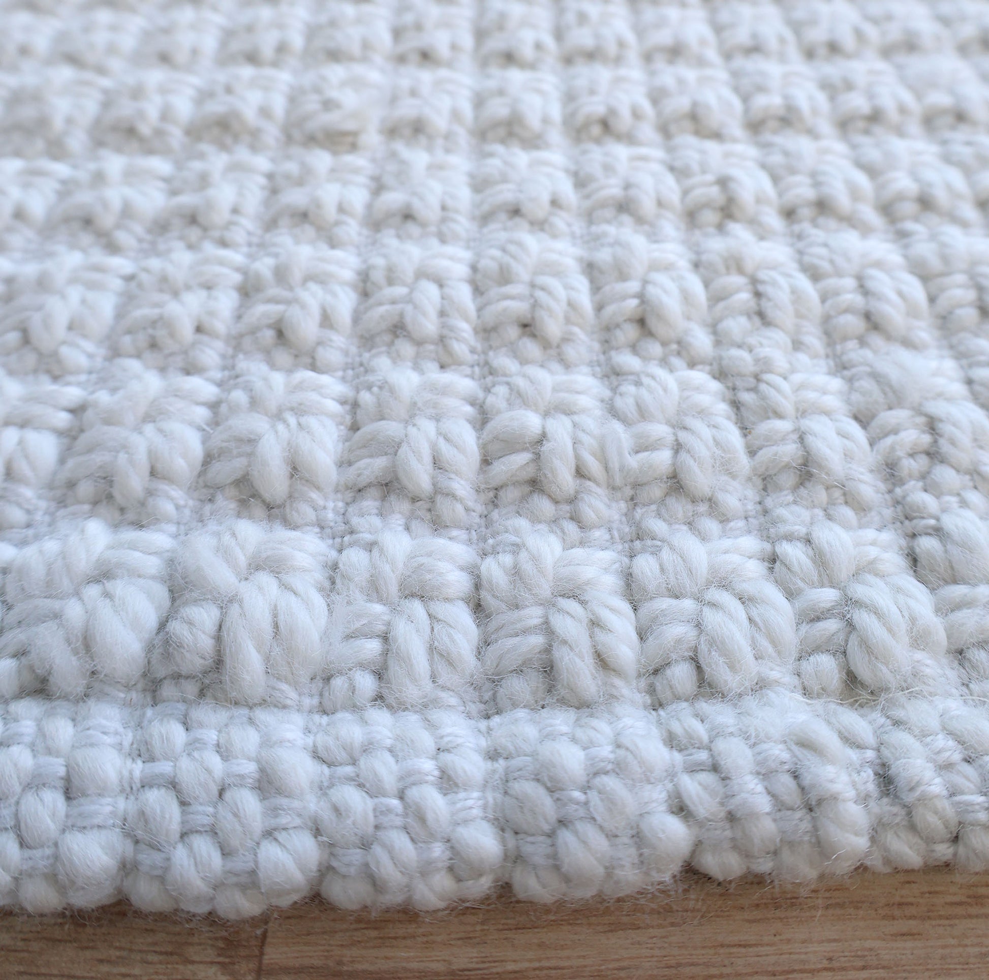 Close-up of the Handmade Cream Natural Color Hand Woven Area Rug WR-039 by RAMSHA HOME, featuring a chunky woven texture and raised, detailed pattern. Laid flat on a wooden surface, this artisanal area rug showcases exquisite craftsmanship and is perfect for those seeking premium home décor crafted from natural fibers.