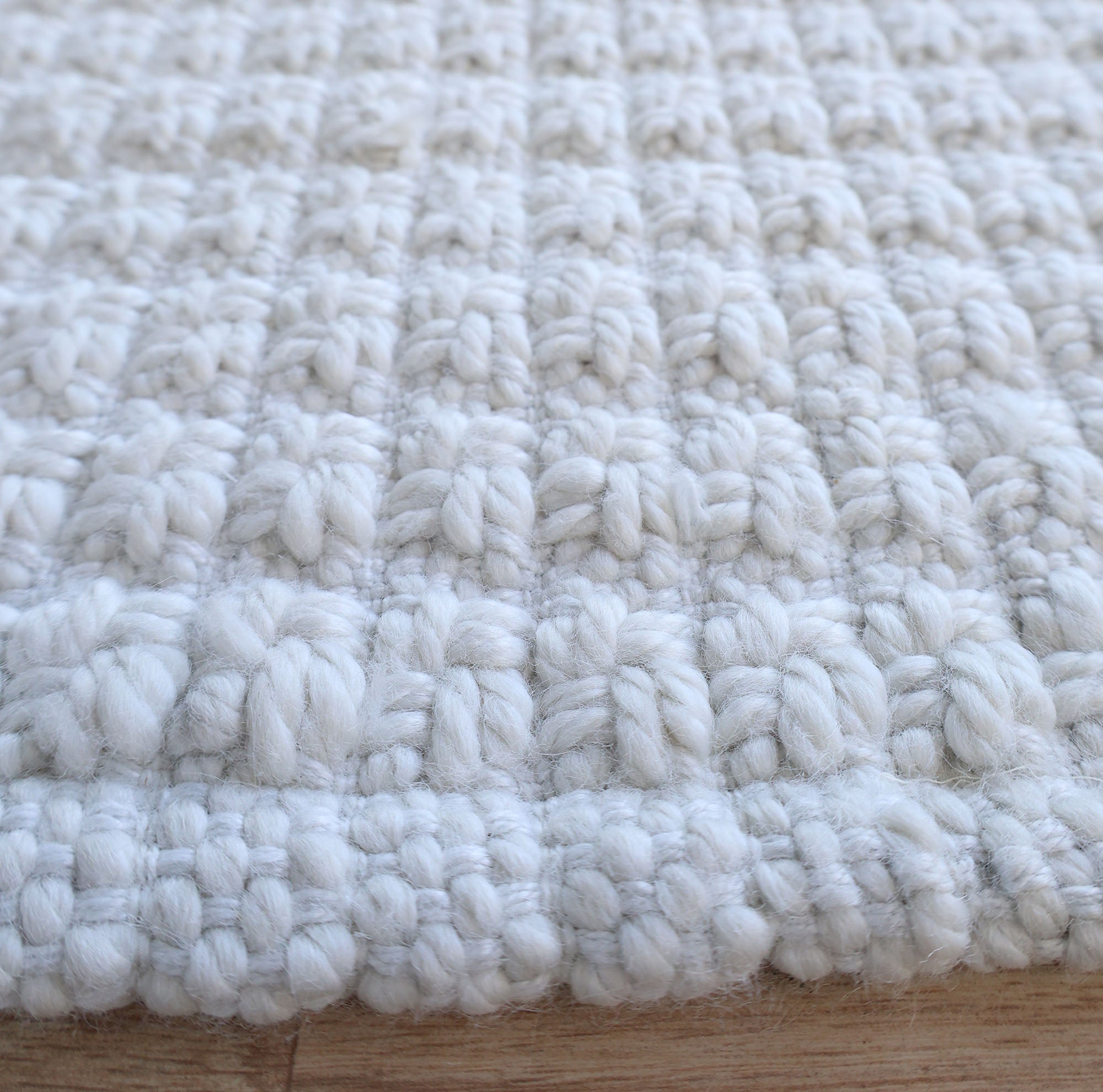 Close-up of the Handmade Cream Natural Color Hand Woven Area Rug WR-039 by RAMSHA HOME, featuring a chunky woven texture and raised, detailed pattern. Laid flat on a wooden surface, this artisanal area rug showcases exquisite craftsmanship and is perfect for those seeking premium home décor crafted from natural fibers.