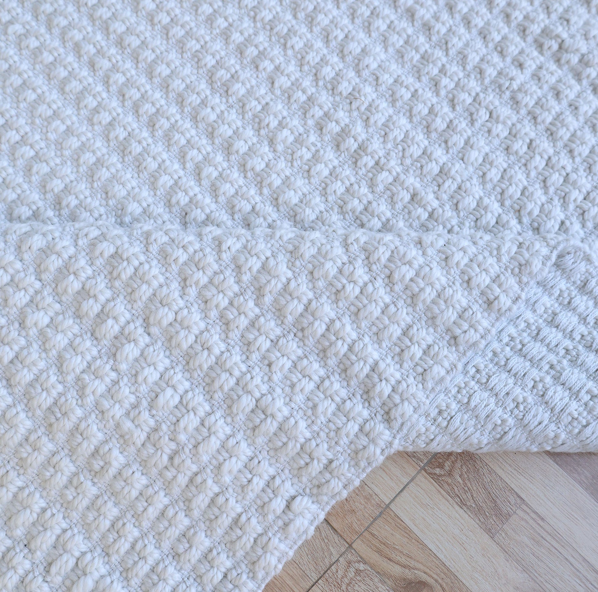 A close-up of the Handmade Cream Natural Color Hand Woven Area Rug WR-039 by RAMSHA HOME, partially folded on a light wooden floor. Crafted from natural fibers, its thick, raised weave adds rich texture and depth—an elegant choice for artisanal home décor.