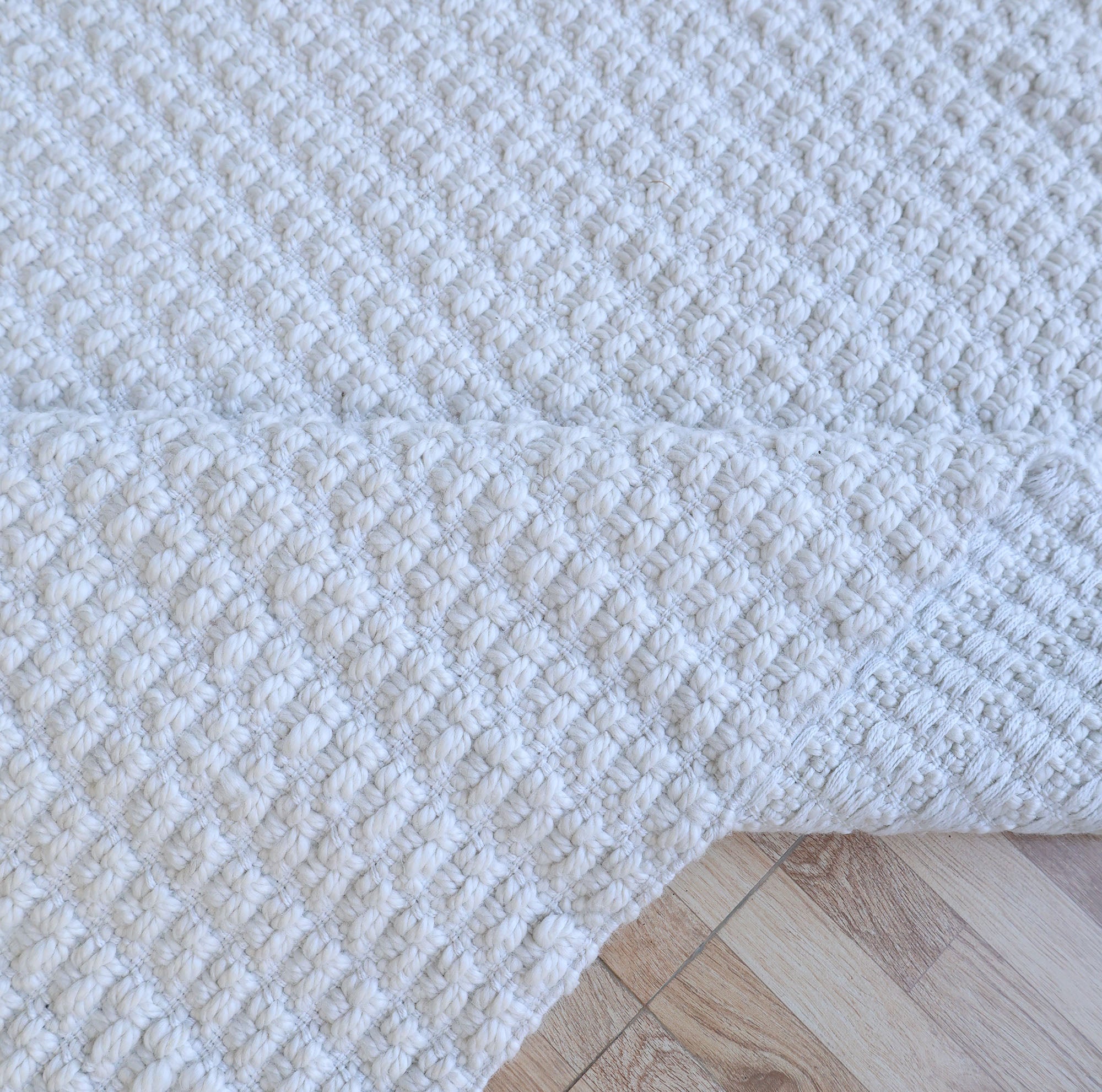 A close-up of the Handmade Cream Natural Color Hand Woven Area Rug WR-039 by RAMSHA HOME, partially folded on a light wooden floor. Crafted from natural fibers, its thick, raised weave adds rich texture and depth—an elegant choice for artisanal home décor.