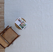 A wooden chair and a small round table with an open magazine are placed on the RAMSHA HOME Handmade Cream Natural Color Hand Woven Area Rug WR-039, set over a wooden floor and viewed from above—an inviting scene that showcases refined artisanal home décor.