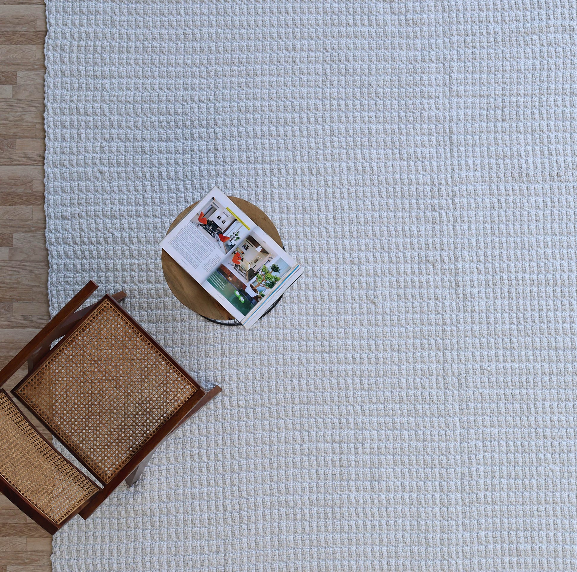 A wooden chair and a small round table with an open magazine are placed on the RAMSHA HOME Handmade Cream Natural Color Hand Woven Area Rug WR-039, set over a wooden floor and viewed from above—an inviting scene that showcases refined artisanal home décor.