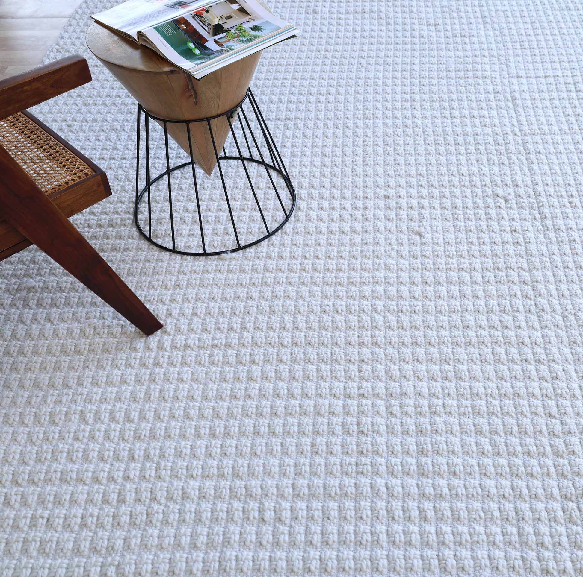 The Handmade Cream Natural Color Hand Woven Area Rug WR-039 by RAMSHA HOME lies beside a wooden chair and a small round side table with a magazine on top, bringing artisanal home décor charm to the space.