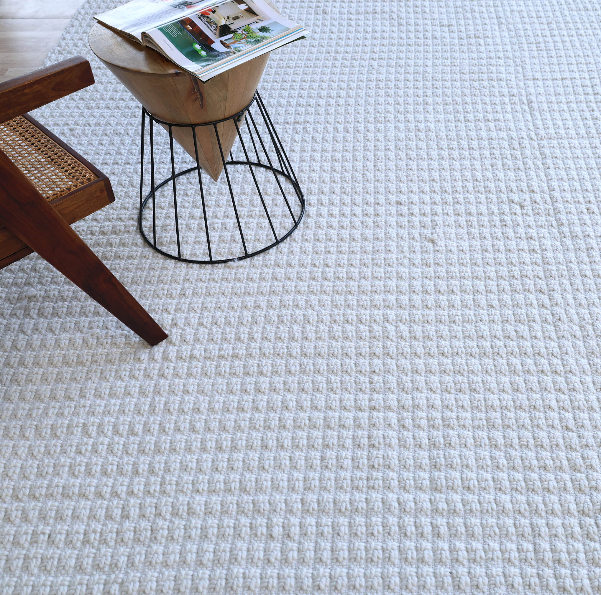 The Handmade Cream Natural Color Hand Woven Area Rug WR-039 by RAMSHA HOME lies beside a wooden chair and a small round side table with a magazine on top, bringing artisanal home décor charm to the space.
