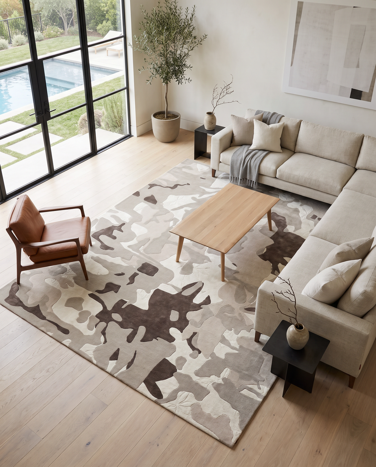 A modern living room featuring a beige sectional sofa, tan armchair, and wooden coffee table is elevated by the RAMSHA HOME Abstract Wool & Viscose Handmade Carpet 300x400 – HTWV-180, whose striking design adds a contemporary touch. Large windows offer views of the pool and lush greenery outside, while minimalist décor is accentuated with potted plants and wall art.