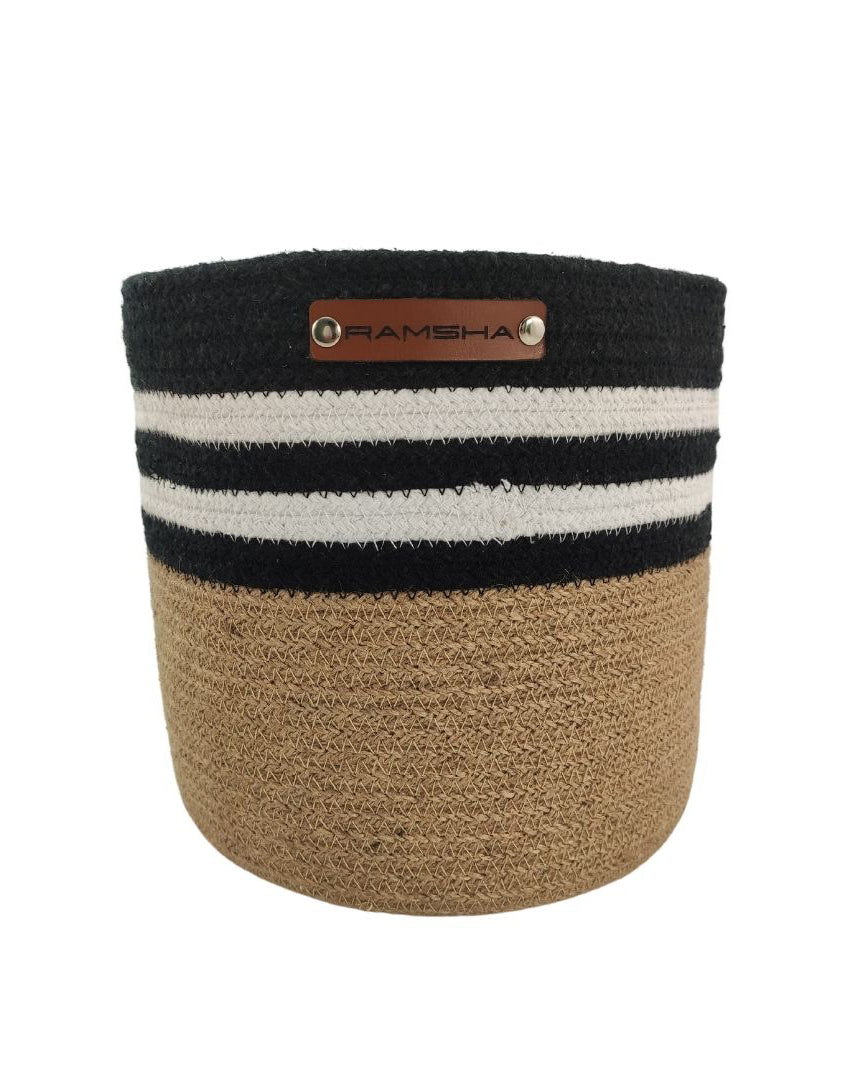 The Jute & Cotton Natural Handmade Basket - BS 004 (25x25cm) by Ramsha Home: Luxury Rugs & Home Furnishings Brand In The UAE is perfect for nursery decor and features a stylish design with the brand's metal tag on the front.