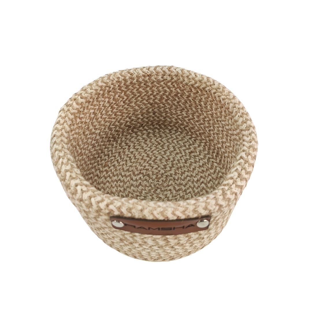 The Ramsha Home Natural and White Handmade Jute and Cotton Basket (BS 008, 12x20 cm) is a small, round decor piece featuring a herringbone pattern and brown label, perfect for adding luxury style to your UAE home.