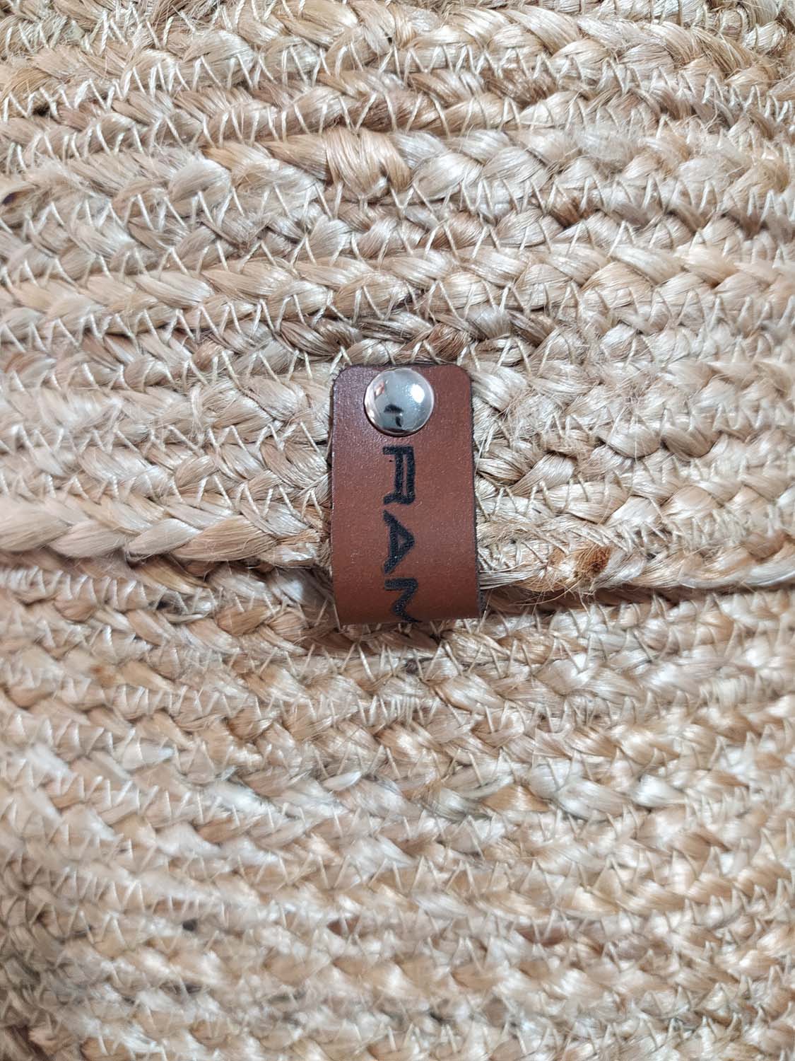 Close-up of the Natural Handmade Jute Basket - BS 016 (20x30cm) by Ramsha Home, showing woven straw texture and a brown leather tag with black text—perfect for natural decor or eco-friendly storage.