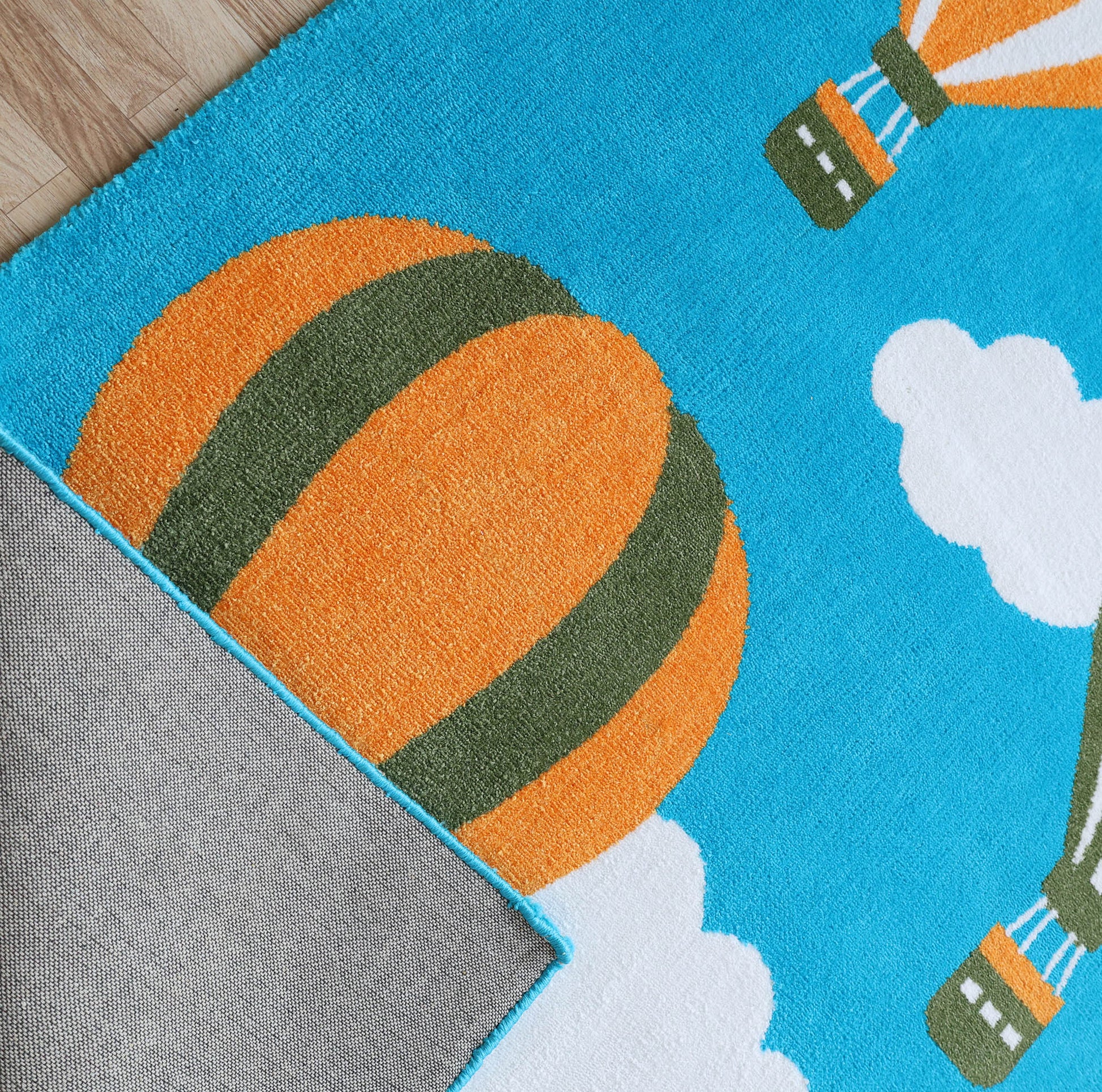 The RAMSHA HOME Kids Play Rug with Clouds & Balloons (CMN 101, 200x300 cm) features an orange and green balloon on a blue background with white clouds. One corner is flipped, showing the gray underside and floor beneath—perfect for a nursery.