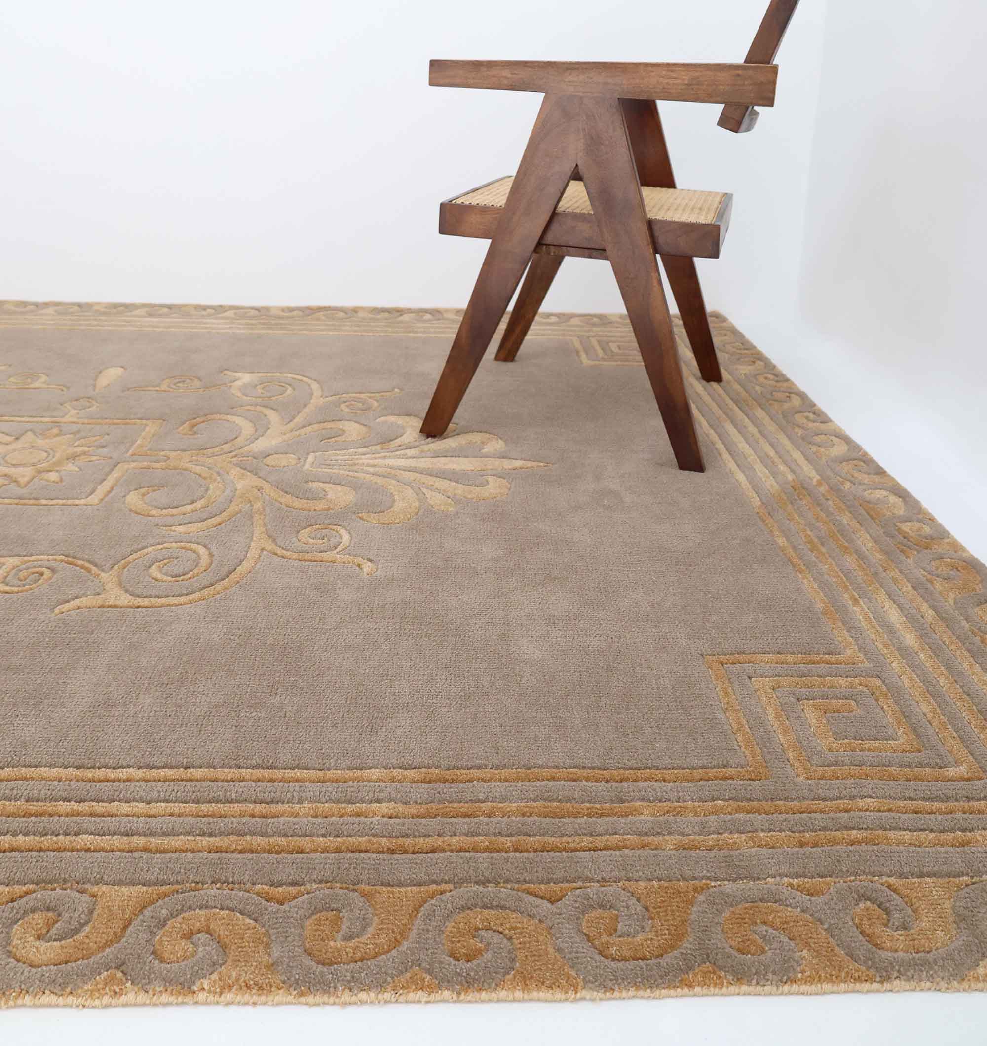 A wooden chair with angled legs sits on a RAMSHA HOME Beige Rectangle Novara Rug (CMN 22, 200x300cm), which features beige and gold geometric and swirling patterns, all set against a plain white background.