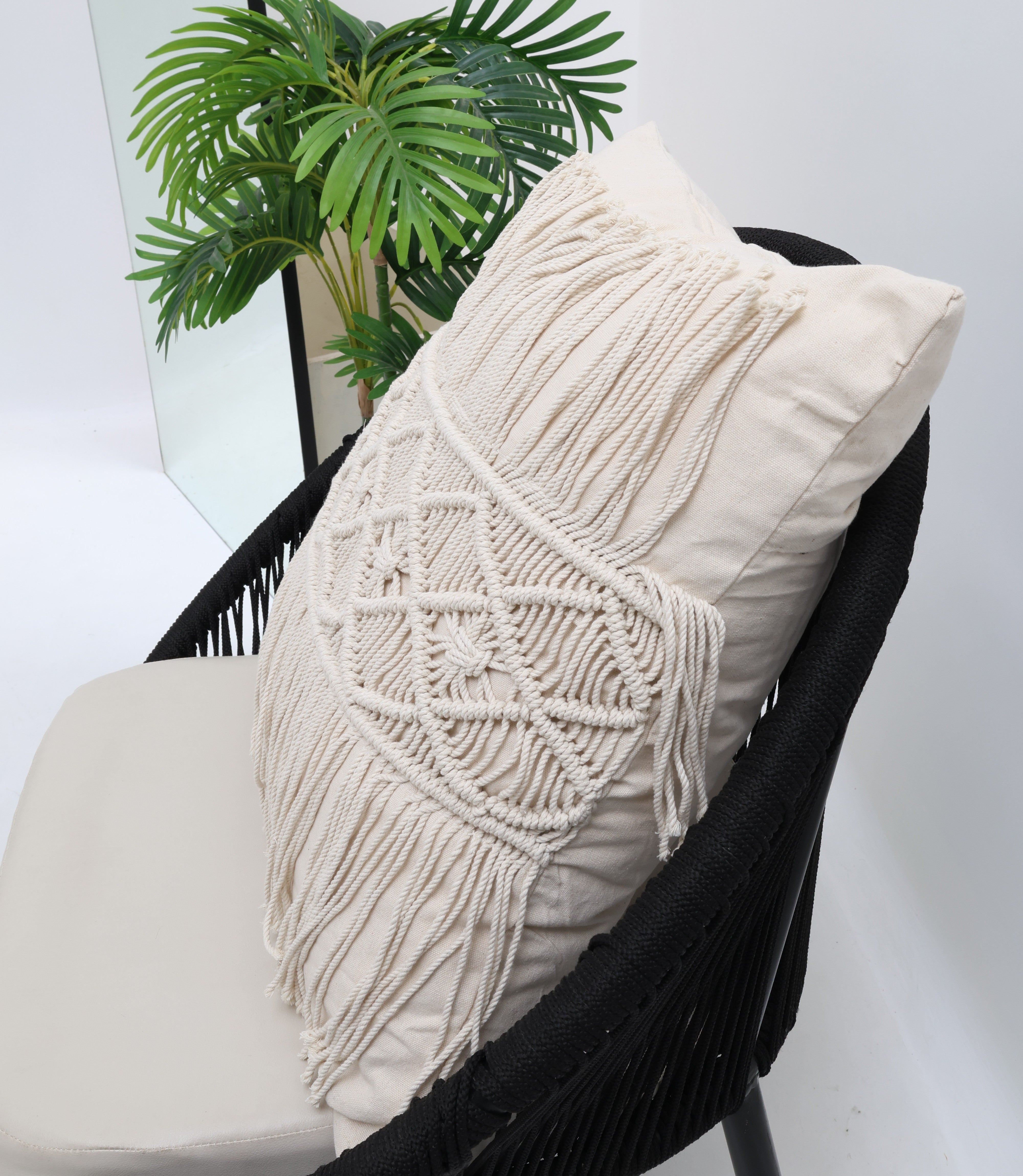 A black chair with Ramsha Home’s Natural Color Jute Cushion with Filler (CS 307, 45x45cm) and a decorative macramé pillow sits beside a green potted plant in a bright, minimalistic room.