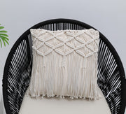 The Ramsha Home Natural White Macrame Sofa Cushion (CS 309), featuring intricate woven patterns and fringe, rests on a black rounded chair with a light cushion; a green leaf peeks in against a plain white background.
