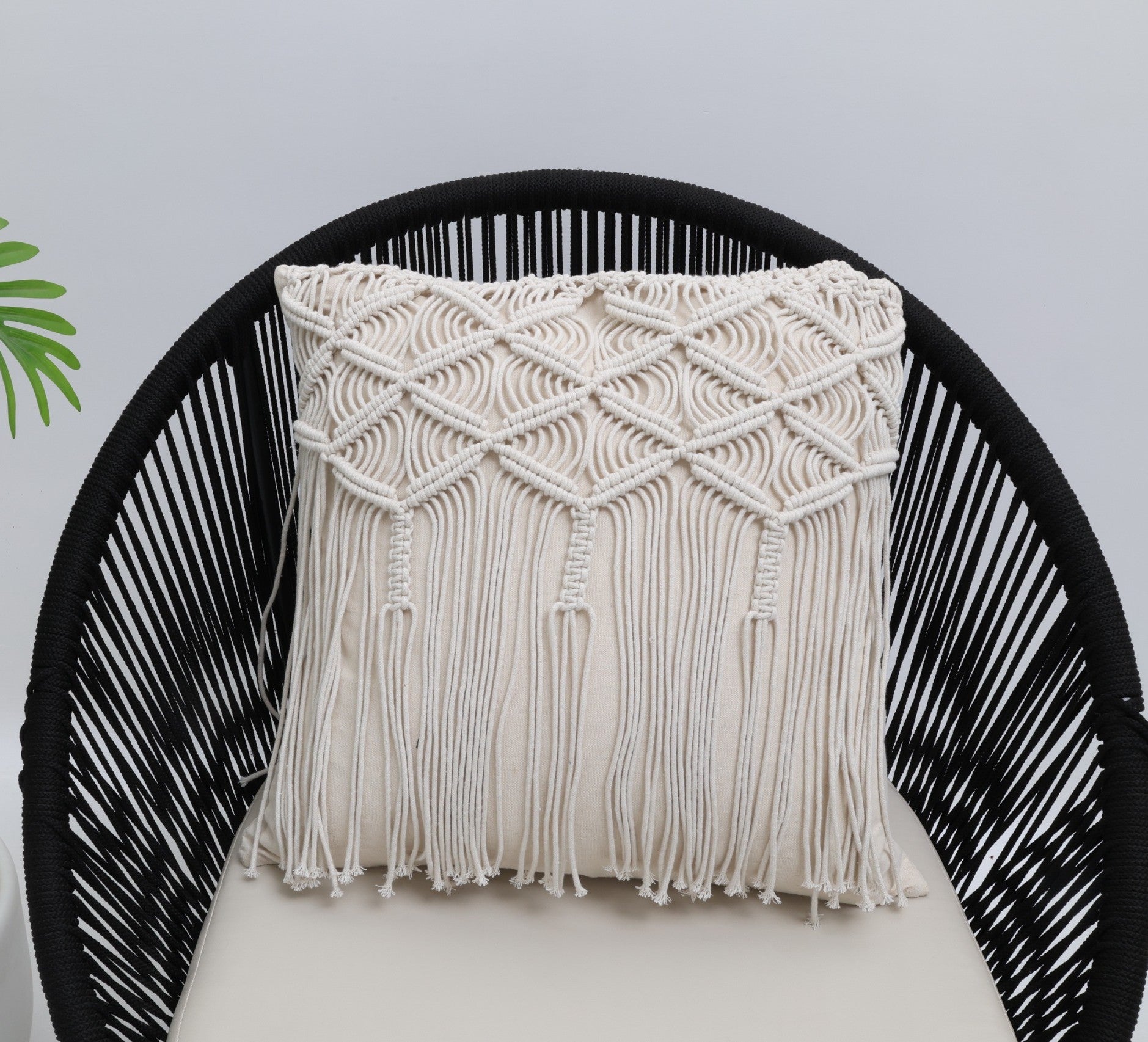 The Ramsha Home Natural White Macrame Sofa Cushion (CS 309), featuring intricate woven patterns and fringe, rests on a black rounded chair with a light cushion; a green leaf peeks in against a plain white background.
