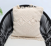 The Natural White Macrame Cushion - CS 311 by Ramsha Home rests on a black woven chair with a light cushion, while a green plant is partially visible to the left.