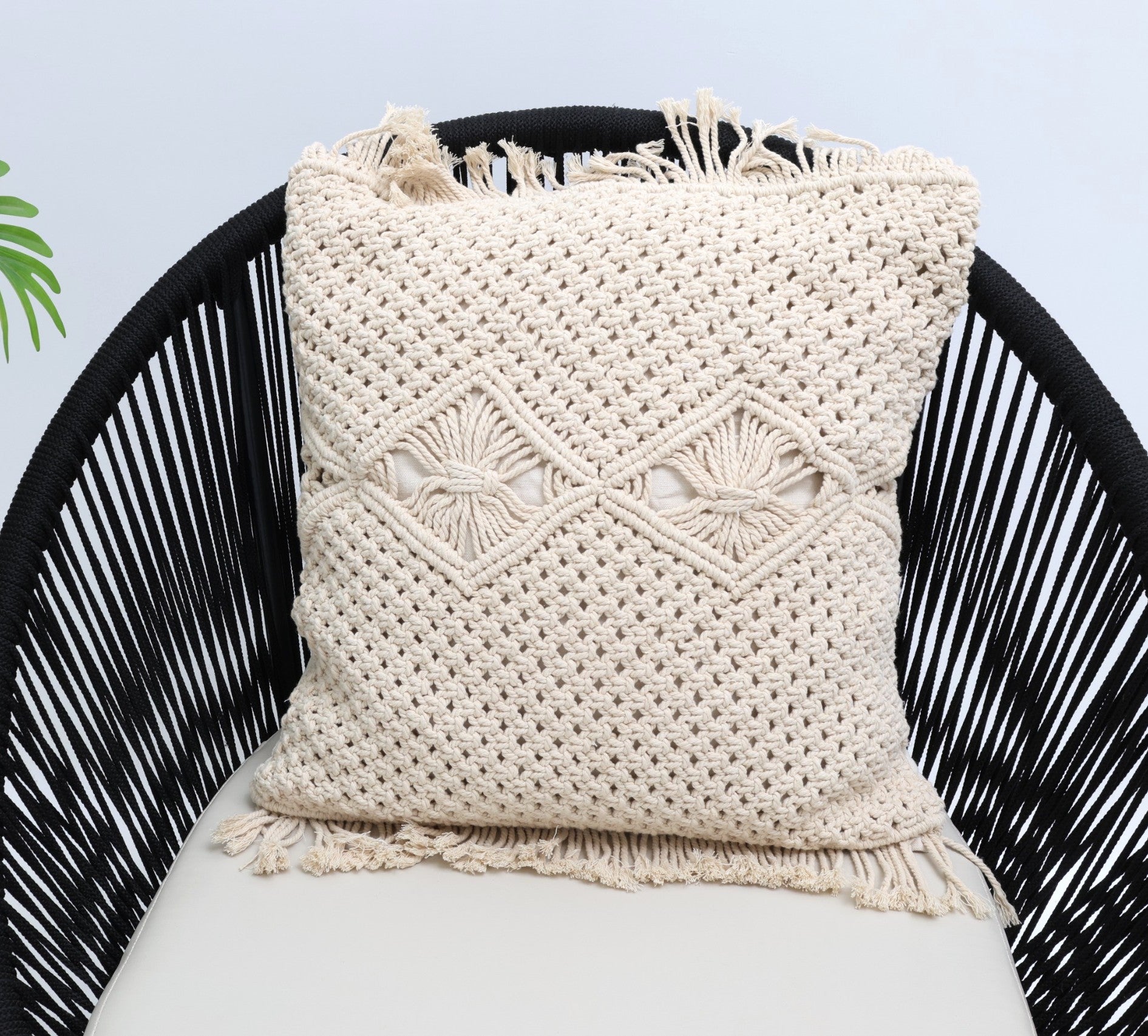 The Natural White Macrame Cushion - CS 311 by Ramsha Home rests on a black woven chair with a light cushion, while a green plant is partially visible to the left.